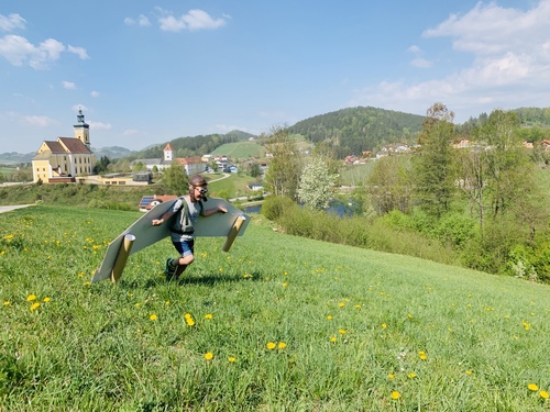 Ein Junge mit Fliegermütze spielt auf einer Wiese mit selbst gebastelter Tragfläche und träumt vom Fliegen über den Wolken. Ringsum Felder, Bäume und eine Kirche